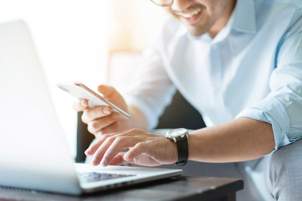 photo of a smiling man using his computer and cell phone